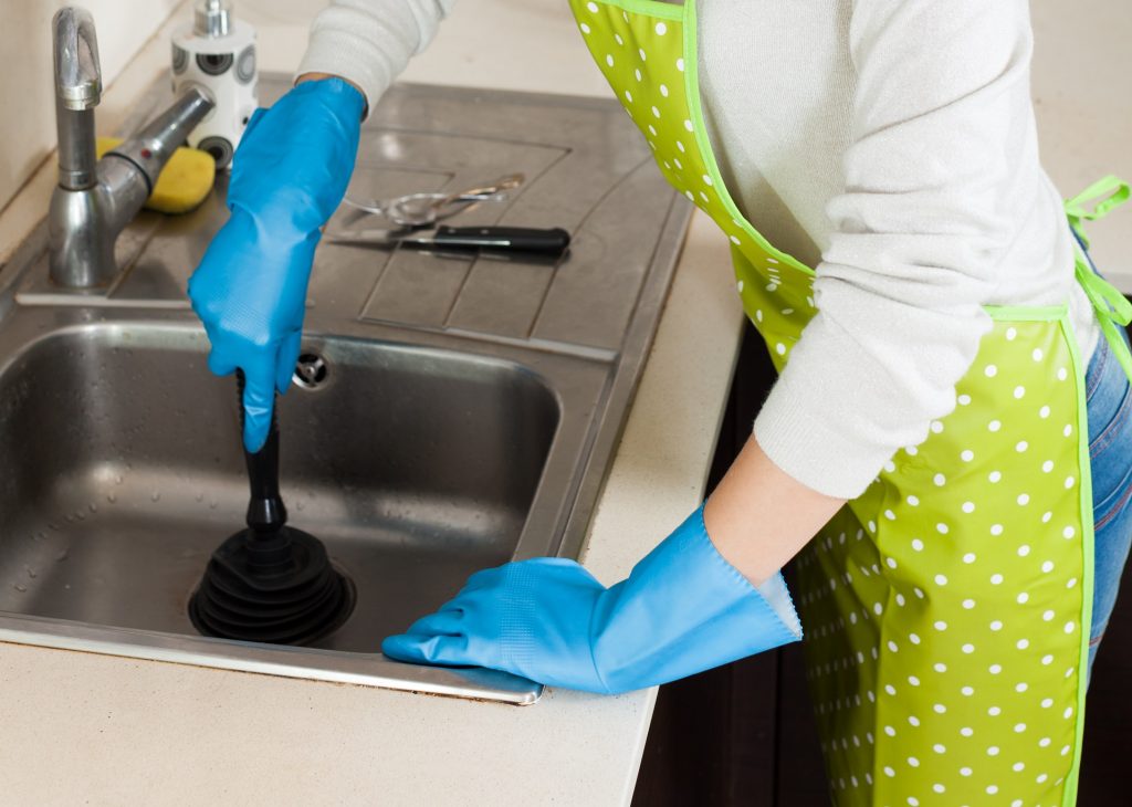 Blonde woman cleaning pipe with cup plunger in kitchen at home